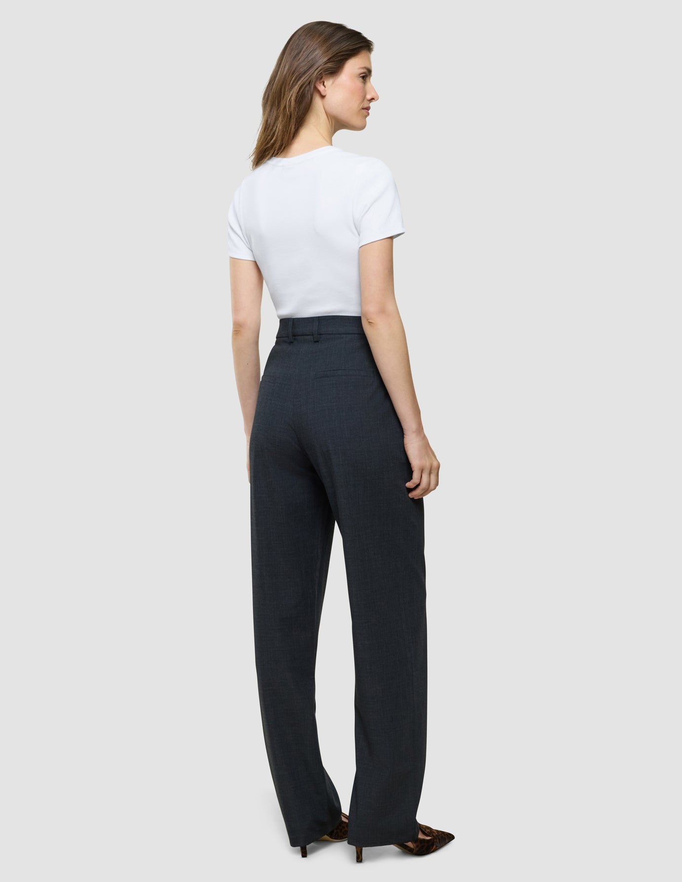 Eine Frau, die mit dem Rücken zur Kamera steht und ein weißes T-Shirt trägt, das in eine hoch taillierte „Women Nara Pants Straight Grey Flint“ gesteckt ist.